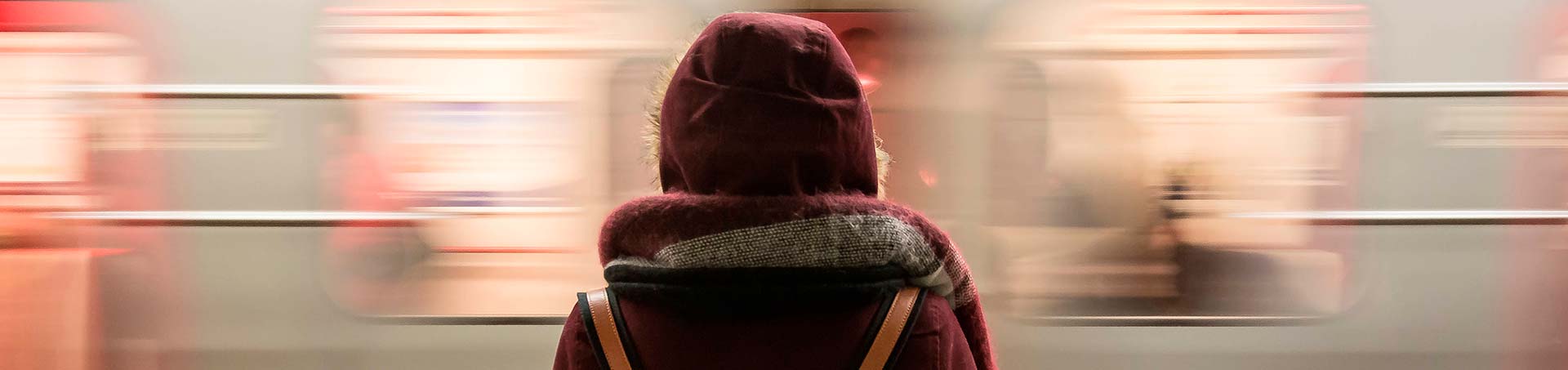 Persona de espaldas, con la capucha puesta, esperando en el andén mientras un tren pasa borroso