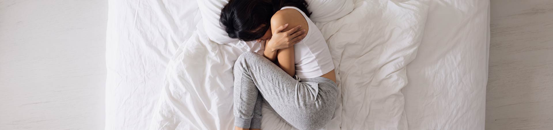 Mujer encogida sobre una cama blanca, vista desde arriba, abrazándose las rodillas con la cara escondida