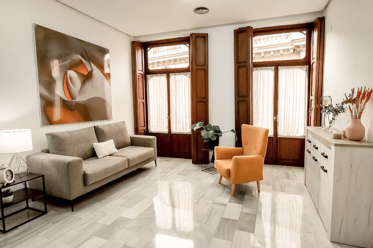 Sala de espera con un sofá, una butaca naranja y dos balcones de madera