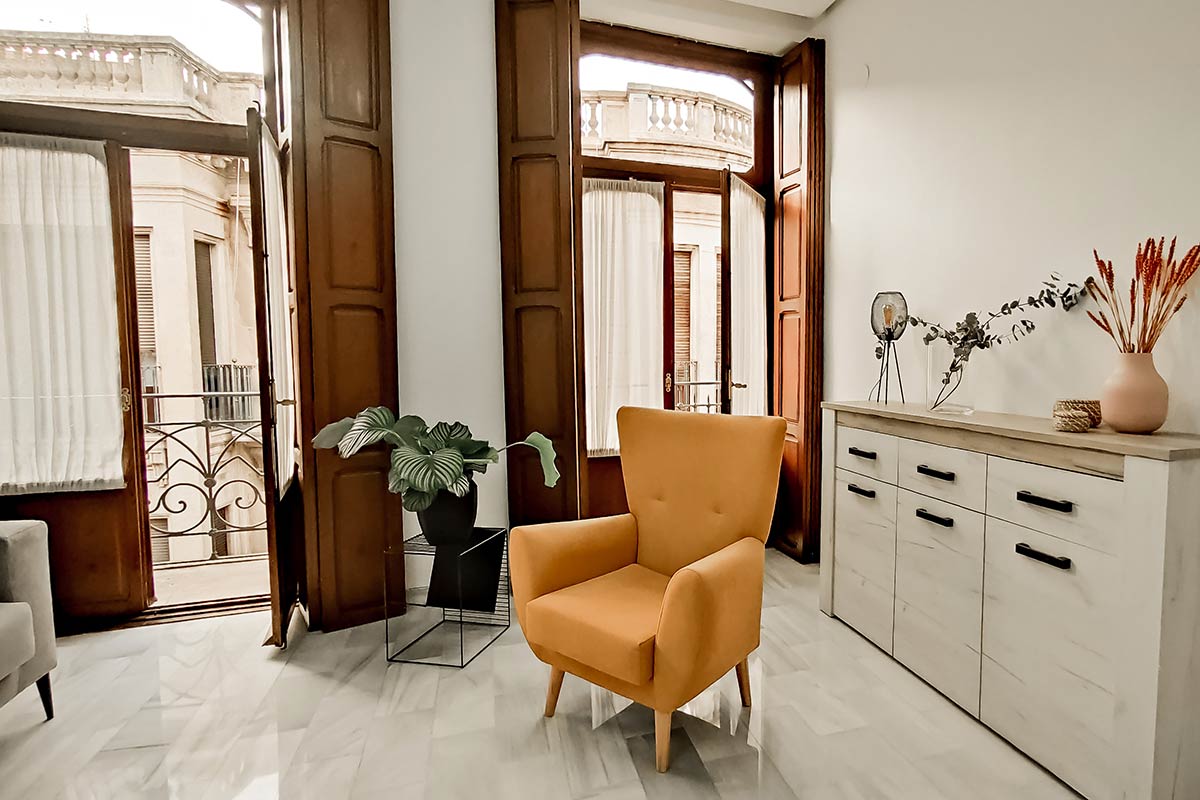 Butaca naranja junto a los balcones abiertos, con una cómoda y plantas