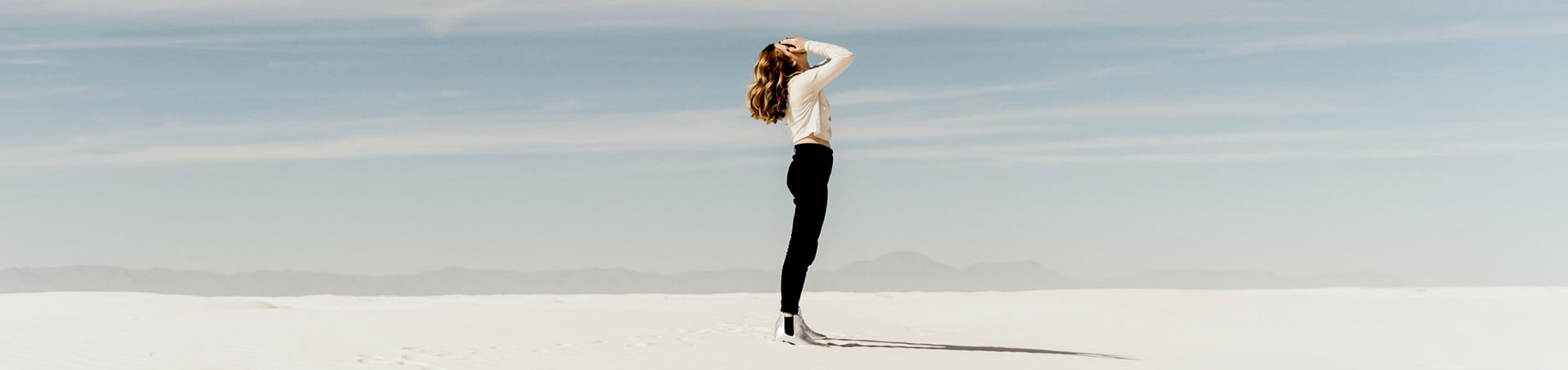 Mujer de pie en medio de un desierto de arena blanca, con las manos en la cabeza y el horizonte despejado